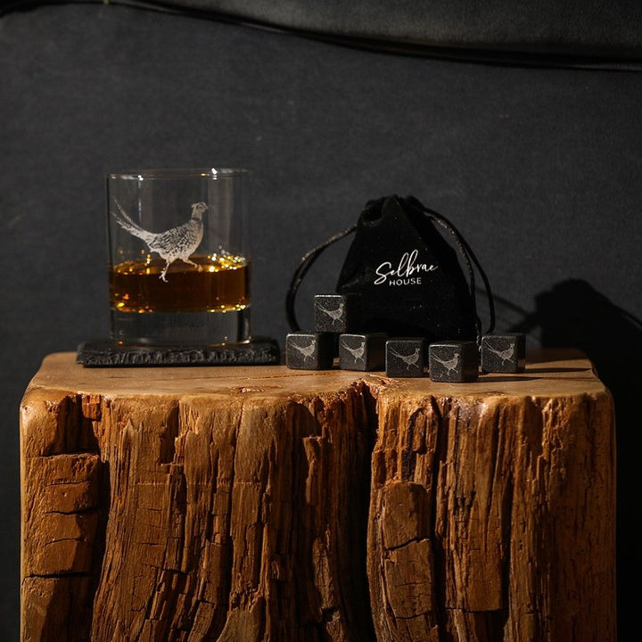 Set of six Selbrae House Pheasant Whisky Stones by Cornucopia Home and Gift, displayed in a clear container on a wooden surface.