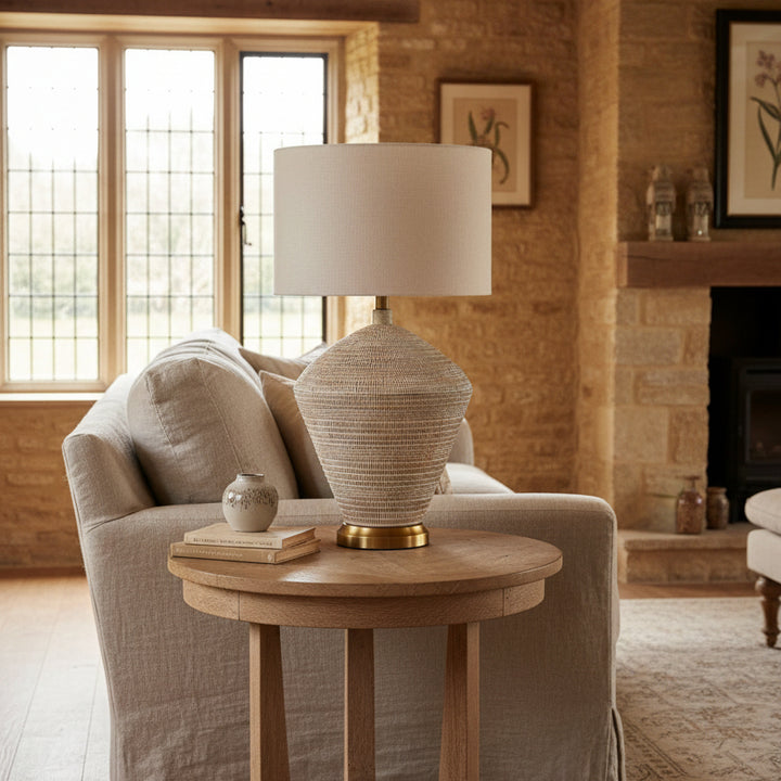 A table lamp with a textured, woven conical base in beige, white, and muted pink tones, topped with a cylindrical beige fabric drum shade and finished with a gold-toned metal base and neck.