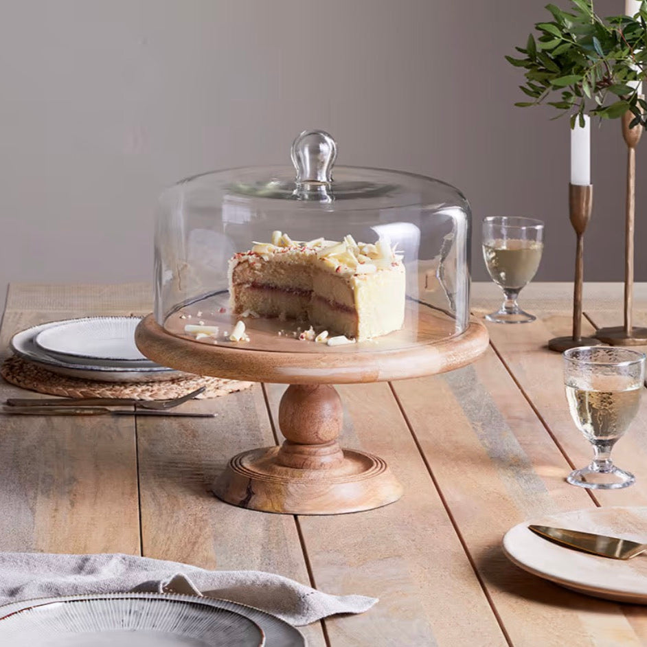 Nkuku recycled glass dome cake stand with clear glass dome and wooden base, ideal for displaying baked goods elegantly.