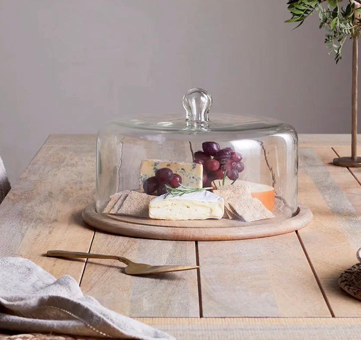 Recycled glass cake dome by Nkuku, showcasing a clear, eco-friendly cover with a rounded shape and wooden base for displaying baked goods.