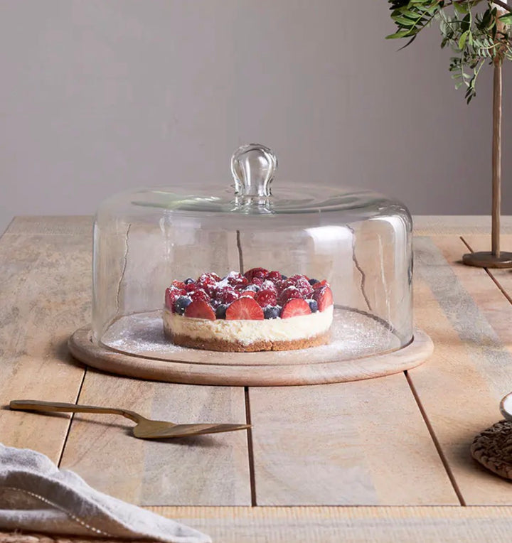 Recycled glass cake dome by Nkuku, clear with a rounded shape and wooden base, displayed on a kitchen countertop.
